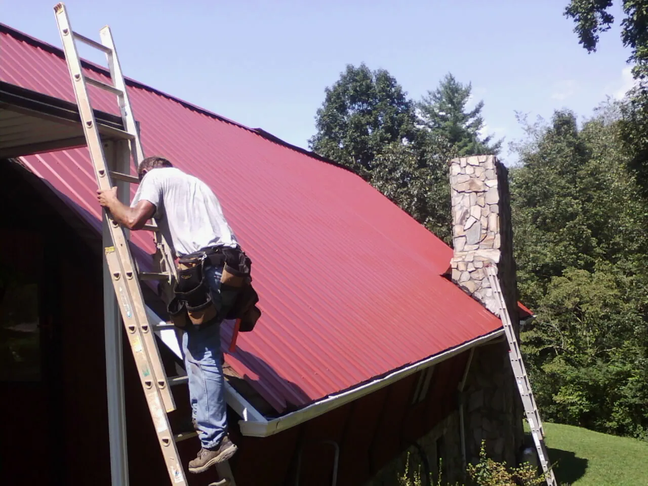 Roofer on a ladder beside a red metal roof and stone chimney — Soffit & Fascia Repair by Roofing Brothers in Battle Mountain